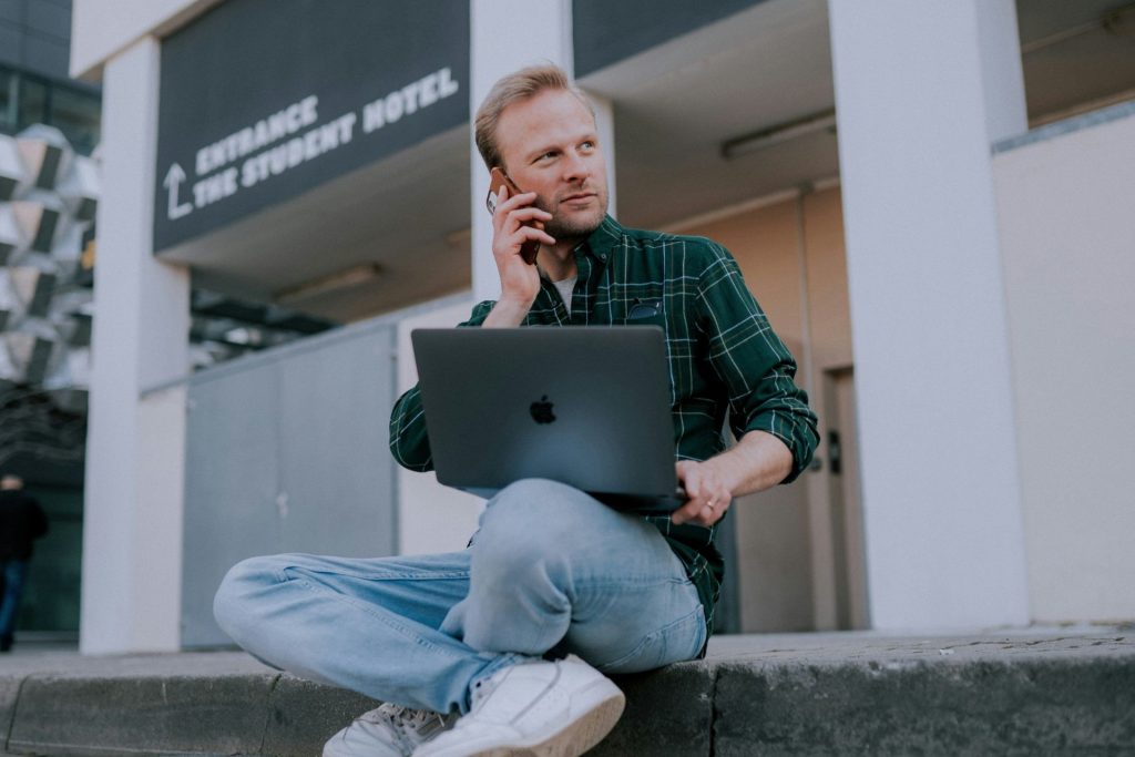 A man sitting on the curb with a laptop and talking on the phone.
