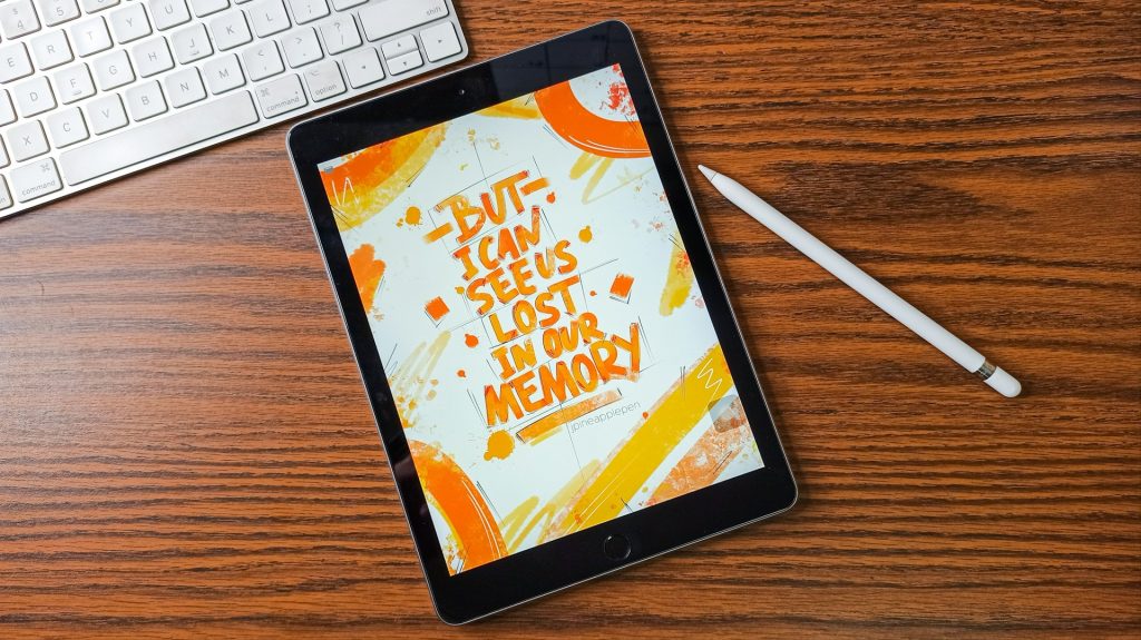 An iPad with Taylor Swift orange and yellow hand drawn text says "But I can see us lost in our memory," which is similar to a Taylor Swift lyric. It's sitting next to an Apple pencil on a wooden surface with a Macbook keyboard above it.