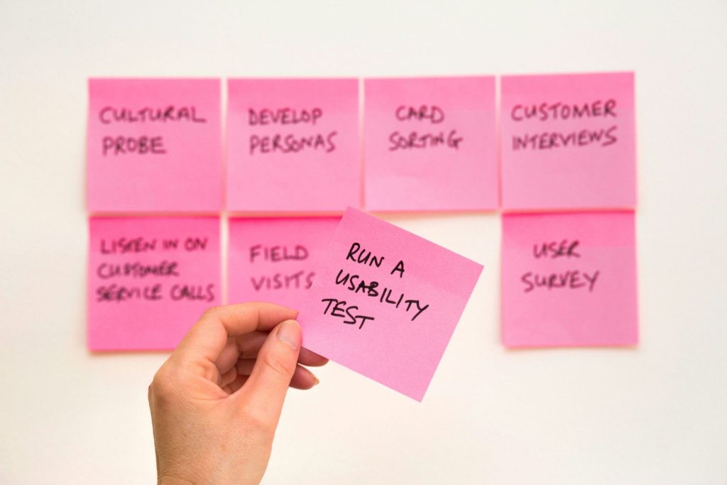 A hand holding a pink sticky note with other sticky notes arranged on a white board labeling various UX research methods.