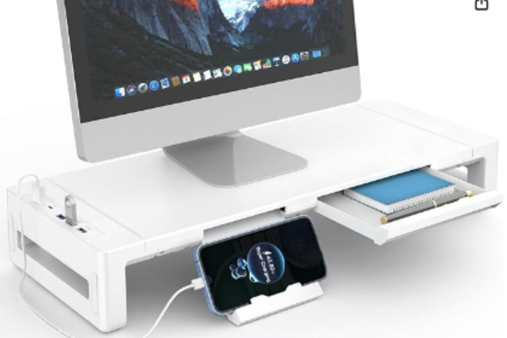 Awhite monitor stand with a blue notebook in storage drawer and phone sitting in stand connected to a charging port.