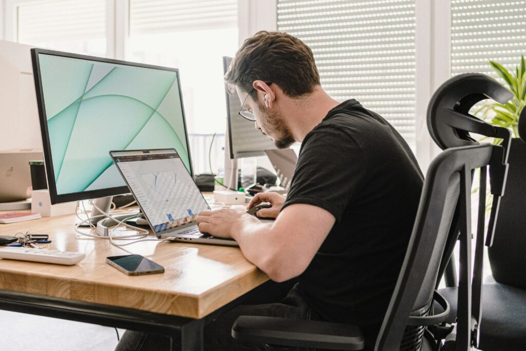 Designer working at a desk with dual monitors and a laptop displaying 3D modeling software