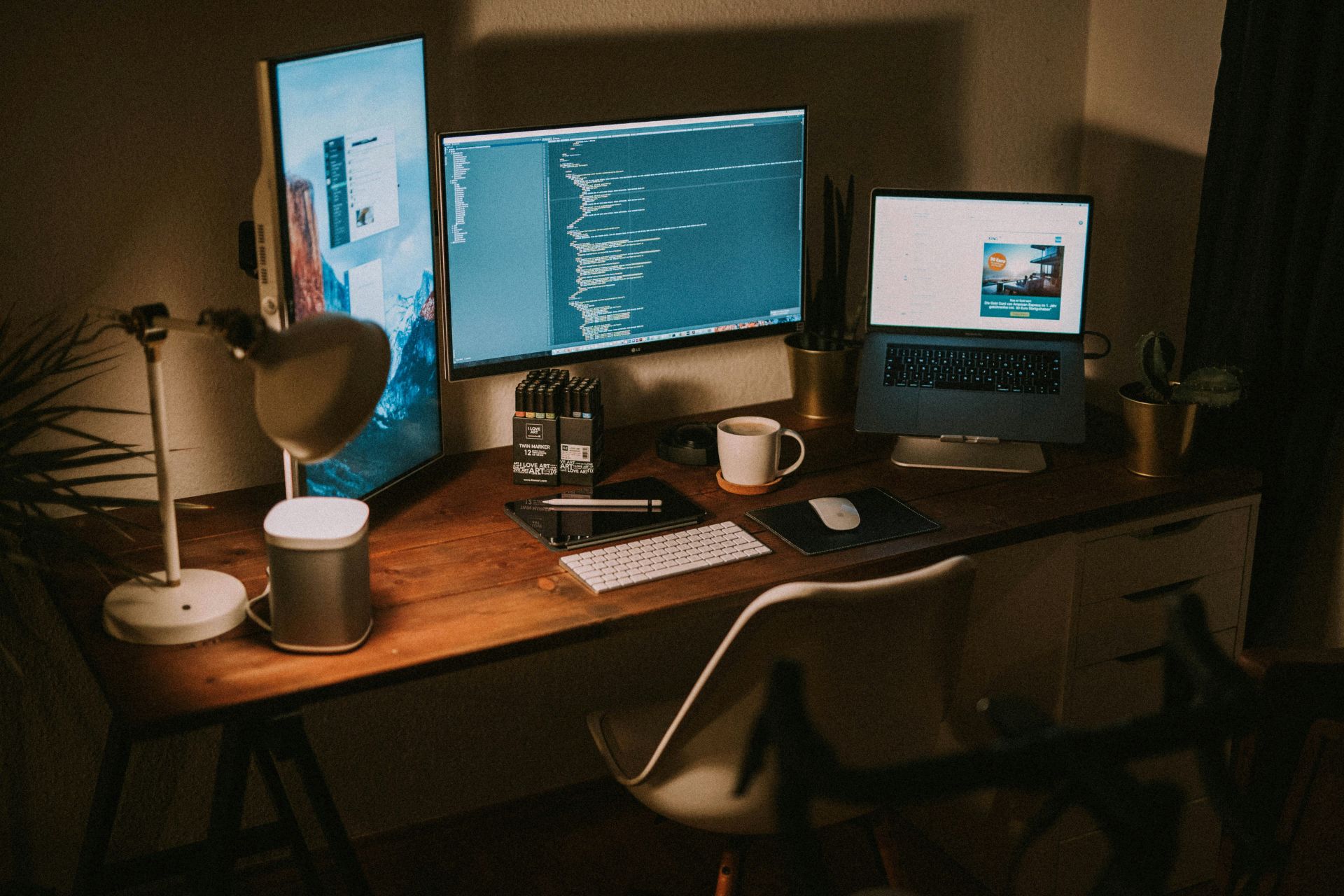 Home workspace with dual monitors dispalying code, a laptop on a stand and desk lamp.