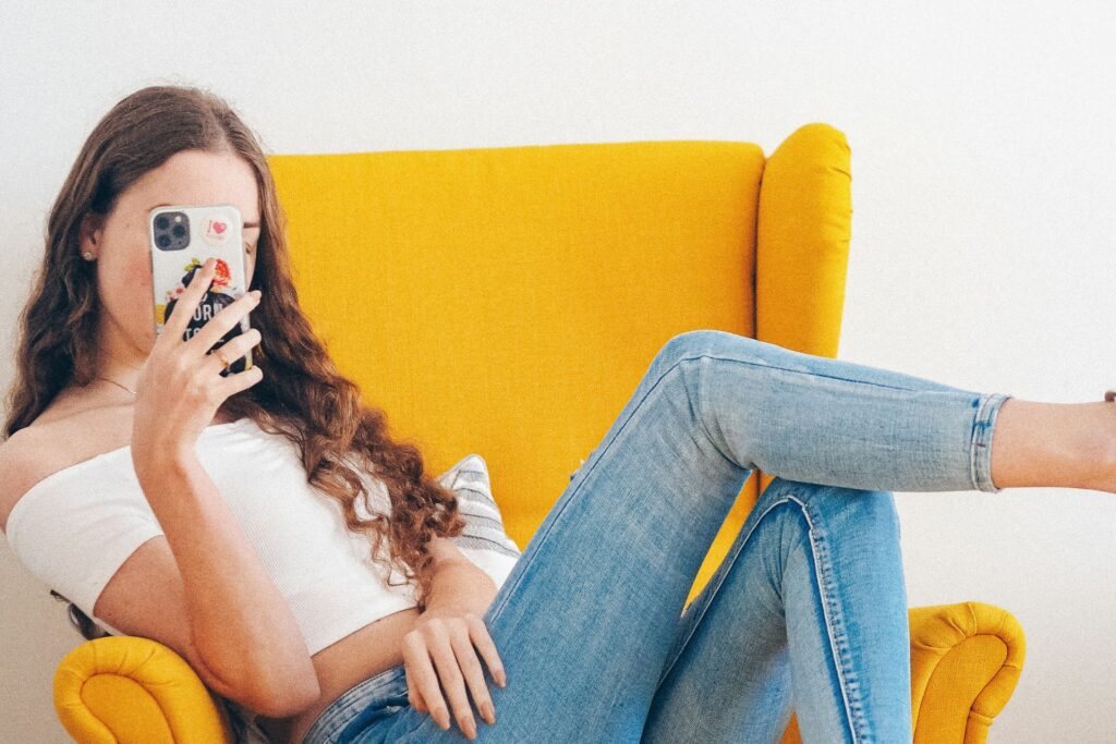 Woman relaxing in yellow chair with feet up while holding a phone that is covering her face.