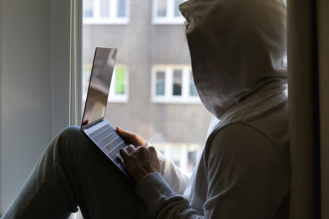 Person wearing a hoodie sitting by a window, using a laptop in a dimly lit area.