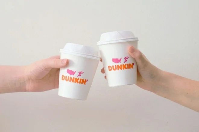 Two hands clinking white paper coffee cups printed with the new DUNKIN' wordmark in orange and pink